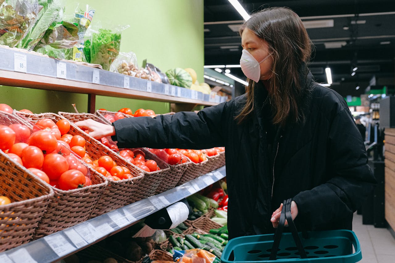 A woman wearing a mask selects fresh tomatoes in a grocery store aisle.